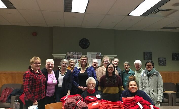 A large group of woman stand and sit together behind many red scarves they have knitted.