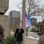 A man speaks into a microphone to a crowd of people outside with the trans pride flag in the background.