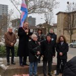 A group of people stand in front of a man with a microphone while he addresses a crowd. The trans pride flies behind them.