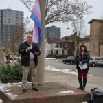 A man with a microphone reads from a book in front of the trans pride flag.