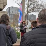 A woman with a microphone addresses a crowd of people. Behind her, the trans pride flag flies.