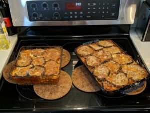 Two baking dishes of cooked eggplant covered in cheese sitting atop a stove.