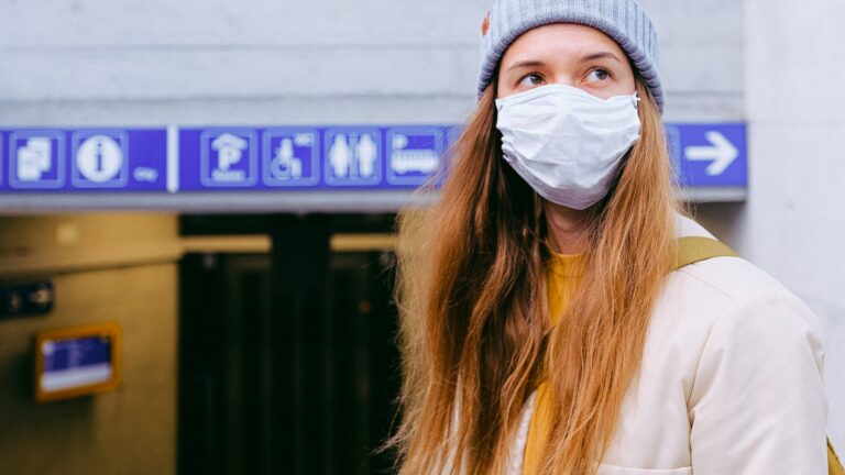 A woman wearing a hat and a medical mask looks off into the distance with a blurred background behind her.