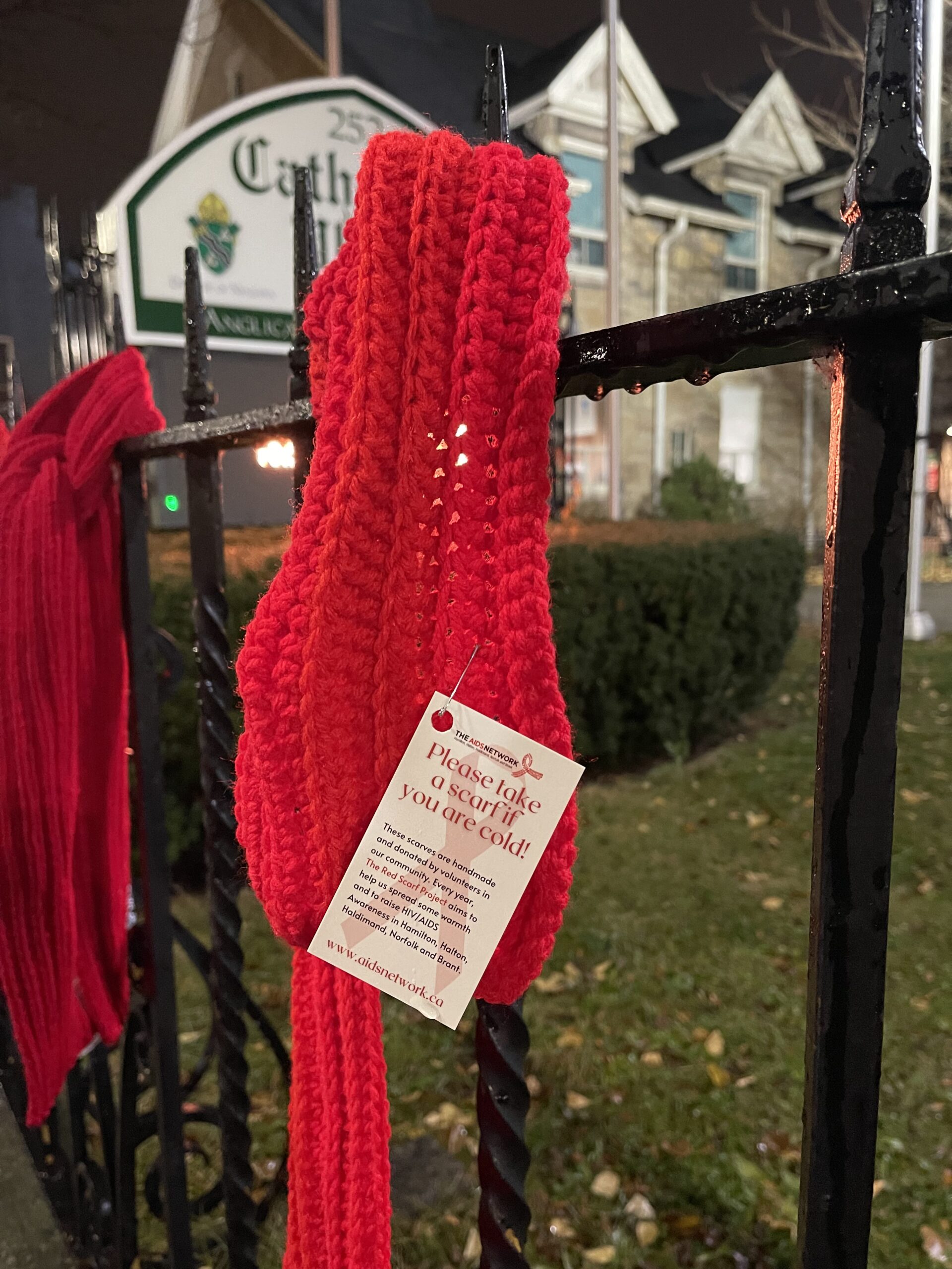 A red scarf hands on a pointed metal fence with a note reading "Please take a scarf if you are cold!"