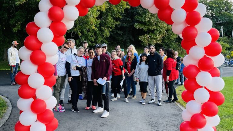 Picture of balloon arch with red and white balloons in Pier 4 Park. A group of walk participants standing behind the arch.